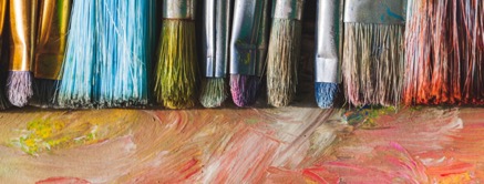 A row of paintbrushes with various colored bristles rests on a wooden surface smeared with colorful paint strokes.