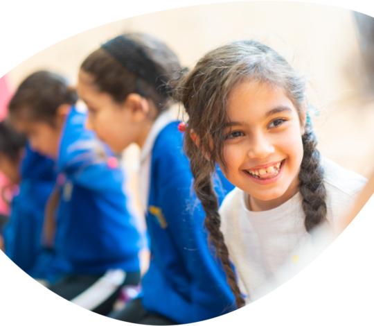 A young girl with braided hair smiles while sitting in a row with other children. They are dressed in blue uniforms.