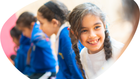 A young girl with braided hair smiles while sitting in a row with other children. They are dressed in blue uniforms.