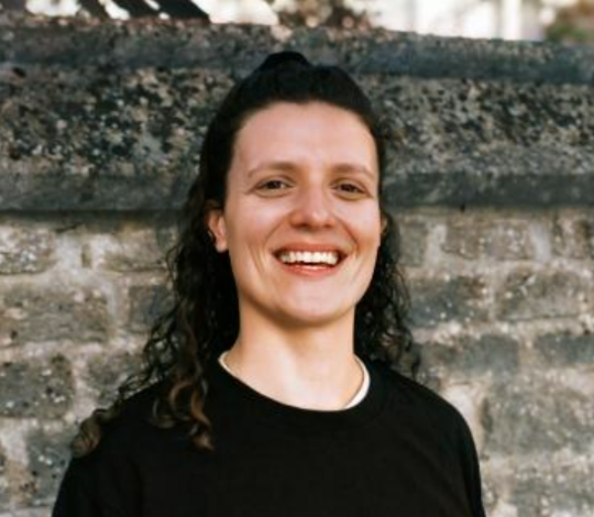 Person with long curly hair smiling and standing in front of a stone wall, wearing a black top.