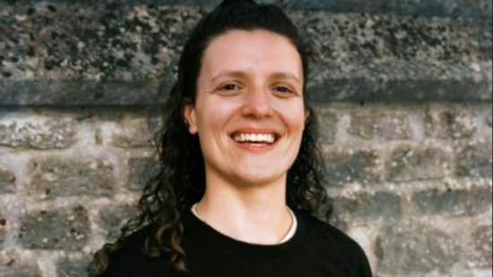 Person with long curly hair smiling and standing in front of a stone wall, wearing a black top.