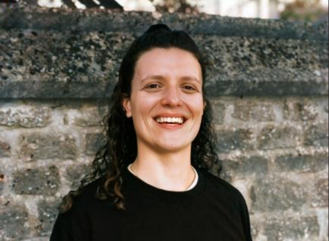 Person with long curly hair smiling and standing in front of a stone wall, wearing a black top.