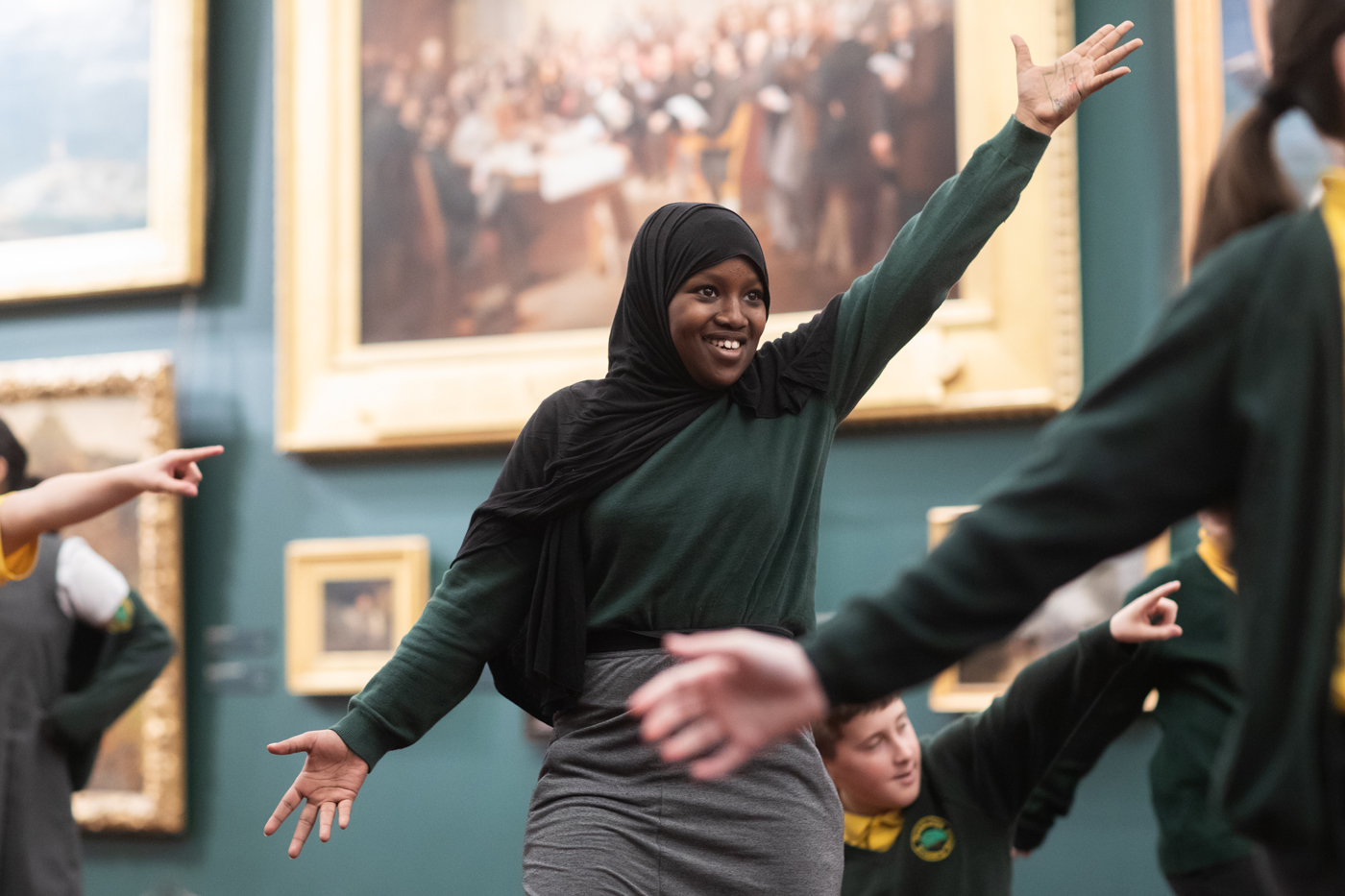 A person wearing a hijab and green sweater poses energetically with arms outstretched in an art gallery, with framed paintings and other individuals in the background.