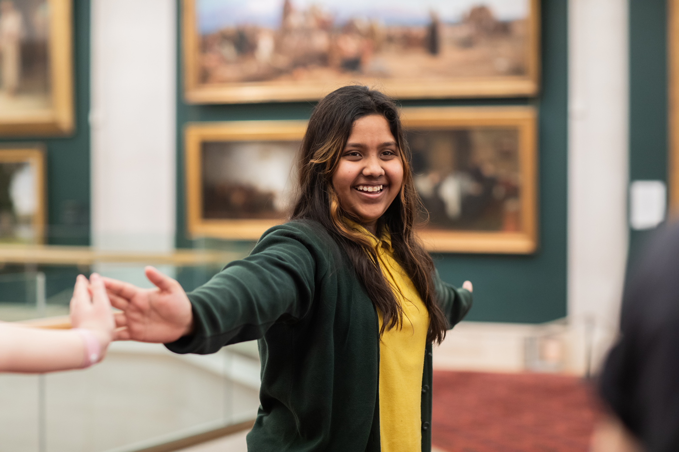 A person smiling with outstretched arms in an art gallery. Paintings in gold frames are visible in the background.