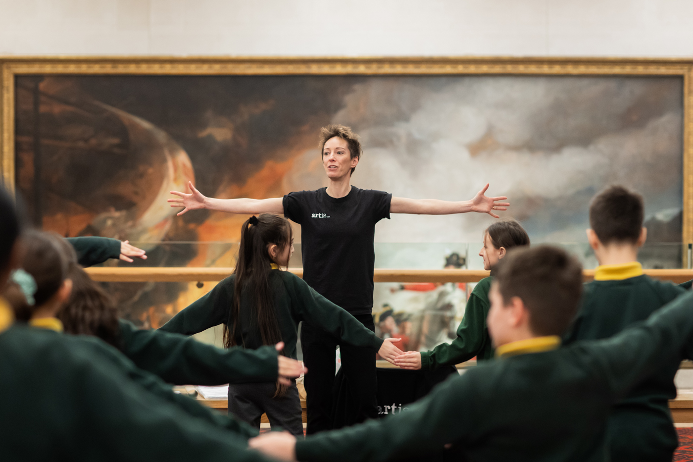 A person in a black shirt guides children in green uniforms through a stretching exercise, with a large painting in the background.