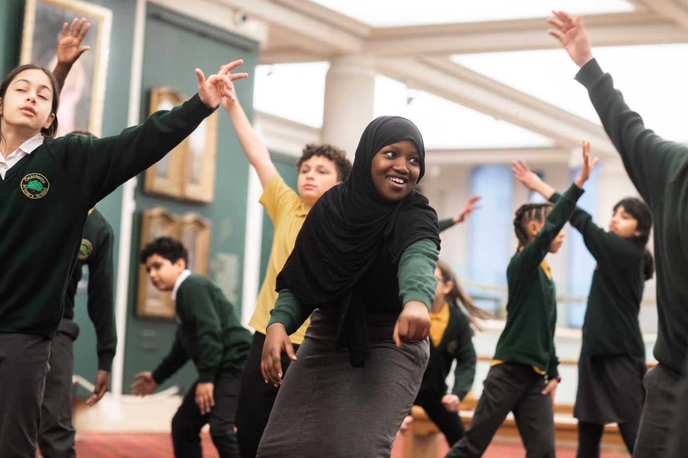 Students participate in a dance or movement activity indoors, smiling and posing with arms raised. They are dressed in school uniforms.