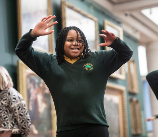 A child with braided hair and wearing a school uniform joyfully raises both hands while standing in an art gallery. A few people are visible in the background.