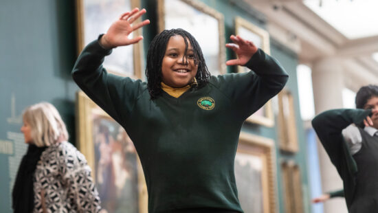 A child with braided hair and wearing a school uniform joyfully raises both hands while standing in an art gallery. A few people are visible in the background.