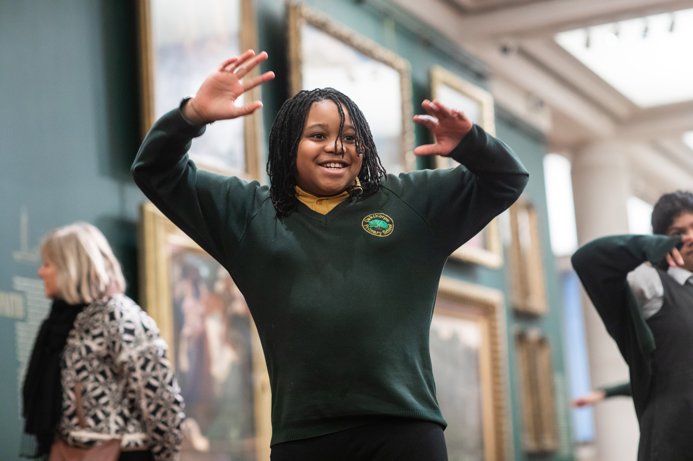 A child with braided hair and wearing a school uniform joyfully raises both hands while standing in an art gallery. A few people are visible in the background.