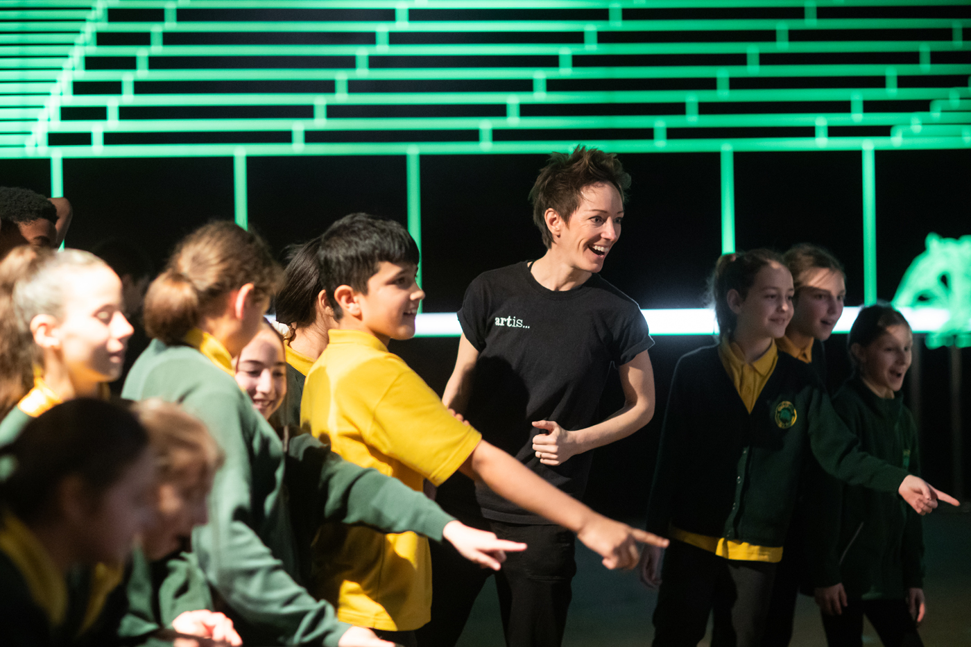 A person in a black T-shirt engages with a group of school children dressed in green and yellow uniforms in a brightly lit room.