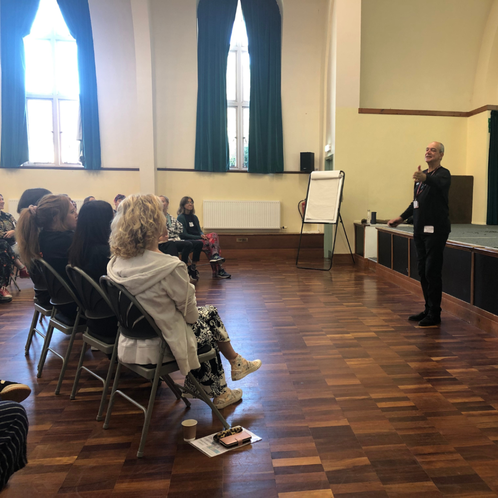 A person is speaking to a seated audience in a room with wooden floors, tall windows with green curtains, and a flipchart.