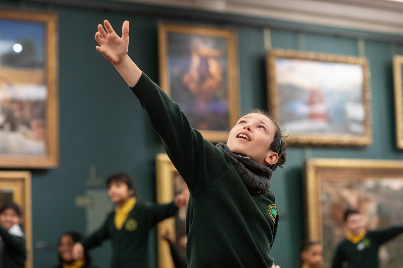 A child in a green uniform joyfully reaches upward, surrounded by classmates, with framed artwork visible in the background.