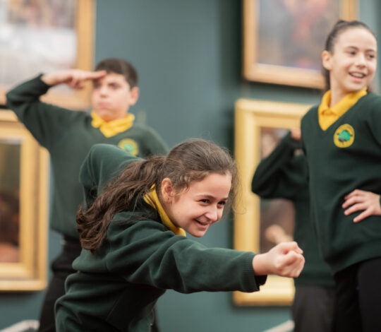 Three children in green school uniforms pose energetically in a room with framed paintings on the wall, one saluting and the others striking lively gestures.