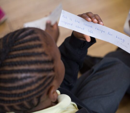 A person with braided hair is holding and reading a handwritten note that says, "Art is the secret recipe for being inspired about life." Another person is partially visible beside them.