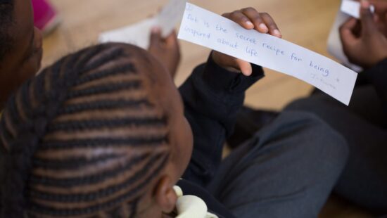 A person with braided hair is holding and reading a handwritten note that says, "Art is the secret recipe for being inspired about life." Another person is partially visible beside them.