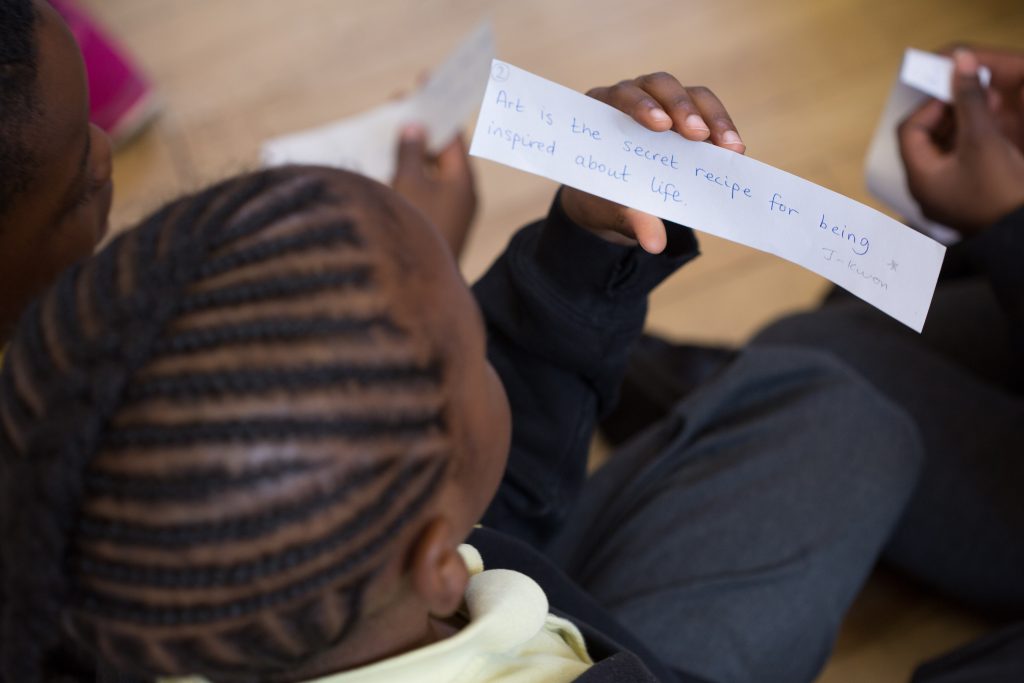 A person with braided hair is holding and reading a handwritten note that says, "Art is the secret recipe for being inspired about life." Another person is partially visible beside them.