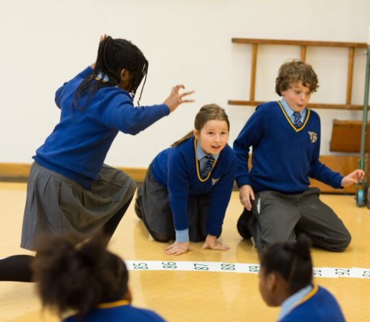 Children in school uniforms participate in an activity on a numbered floor grid in a classroom.