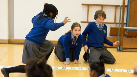 Children in school uniforms participate in an activity on a numbered floor grid in a classroom.