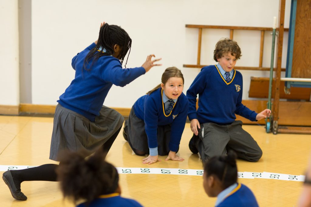 Children in school uniforms participate in an activity on a numbered floor grid in a classroom.