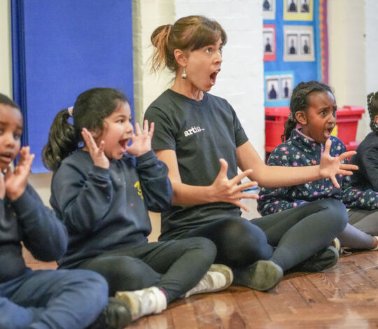 A woman and four children sit on a wooden floor in a classroom, all with their mouths open wide and hands near their faces, appearing to participate in an expressive activity.