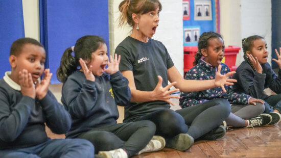 A woman and four children sit on a wooden floor in a classroom, all with their mouths open wide and hands near their faces, appearing to participate in an expressive activity.