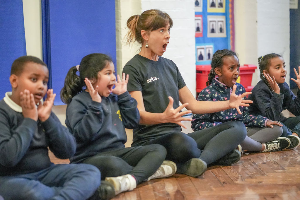 A woman and four children sit on a wooden floor in a classroom, all with their mouths open wide and hands near their faces, appearing to participate in an expressive activity.