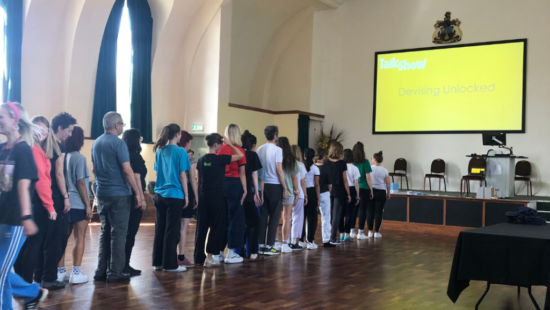 A group of people stands in a line facing forward in a spacious hall with high arched windows, under a large screen displaying the title "Devising Unlocked" as part of a TalkShow event.