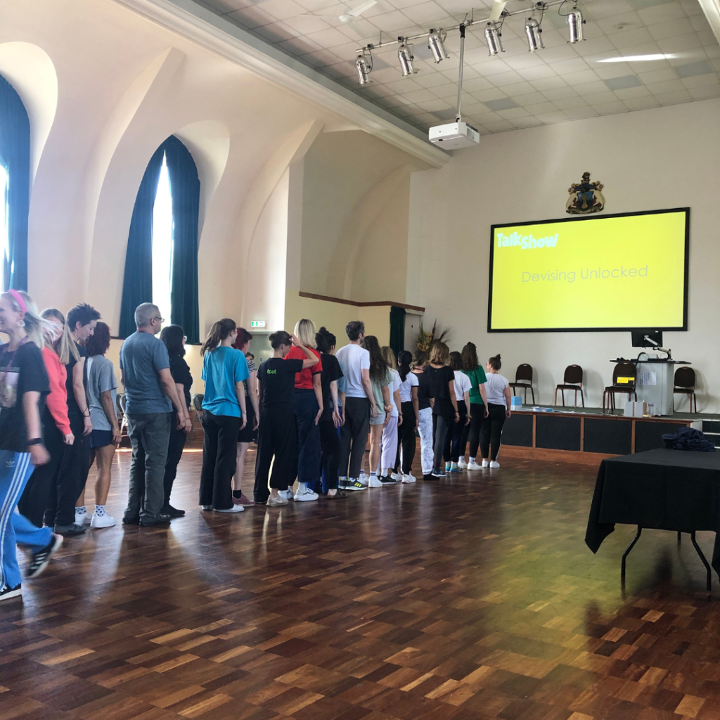 A group of people stands in a line facing forward in a spacious hall with high arched windows, under a large screen displaying the title "Devising Unlocked" as part of a TalkShow event.