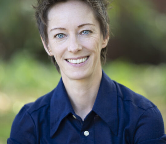 A person with short spiked hair in a navy blue shirt smiles in an outdoor setting with a blurred green and brown background.