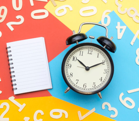 A black alarm clock and a blank notepad are surrounded by scattered wooden numbers on a colorful, divided background.