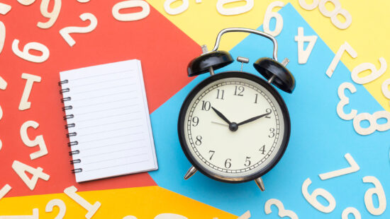 A black alarm clock and a blank notepad are surrounded by scattered wooden numbers on a colorful, divided background.