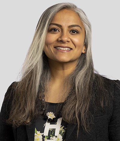 A smiling individual with long, straight, gray and black hair, wearing a black blazer and a floral blouse, poses against a plain gray background.