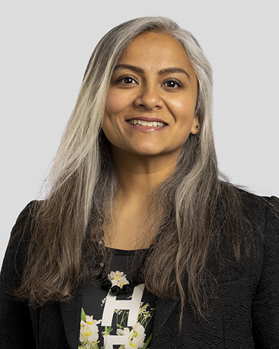 A smiling individual with long, straight, gray and black hair, wearing a black blazer and a floral blouse, poses against a plain gray background.