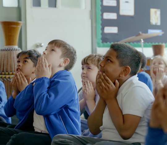 Children in school uniforms sit on the floor in a classroom with their hands held up near their faces, appearing engaged and focused on something out of the frame.