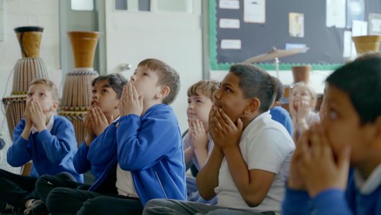 Children in school uniforms sit on the floor in a classroom with their hands held up near their faces, appearing engaged and focused on something out of the frame.