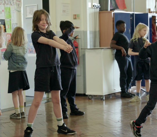 Children in a classroom engage in various activities; one stretches with arms out, another points, and others are standing or moving around. The room has bright decorations and some storage units.