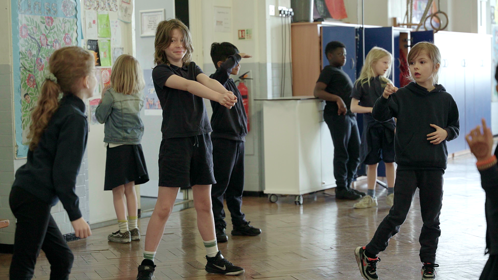 Children in a classroom engage in various activities; one stretches with arms out, another points, and others are standing or moving around. The room has bright decorations and some storage units.