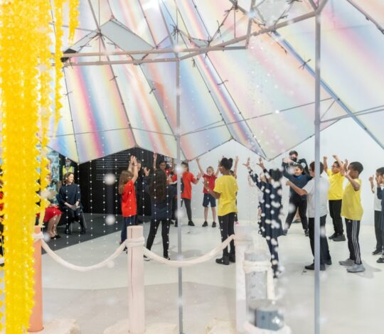 Children participating in a group activity inside a bright, modern room with a dazzling, reflective ceiling and hanging yellow decorations.