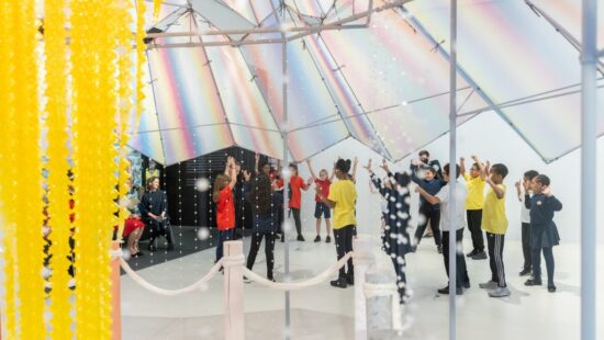 Children participating in a group activity inside a bright, modern room with a dazzling, reflective ceiling and hanging yellow decorations.