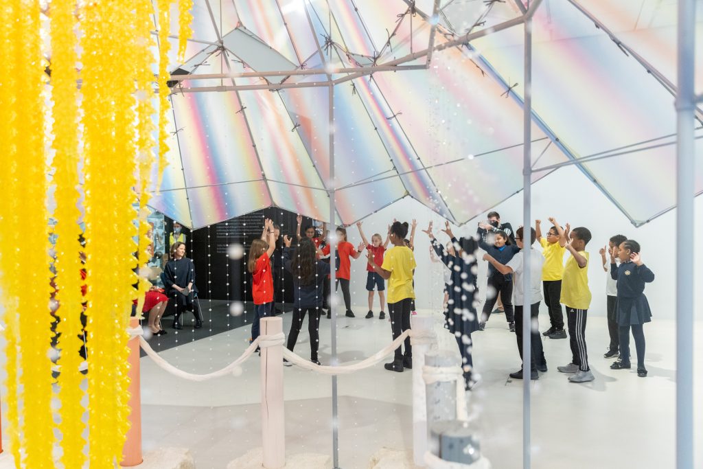 Children participating in a group activity inside a bright, modern room with a dazzling, reflective ceiling and hanging yellow decorations.