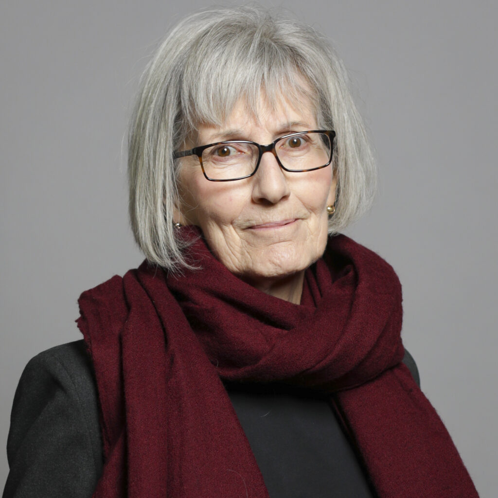 An elderly woman with gray hair wearing glasses and a burgundy scarf stands against a gray background.