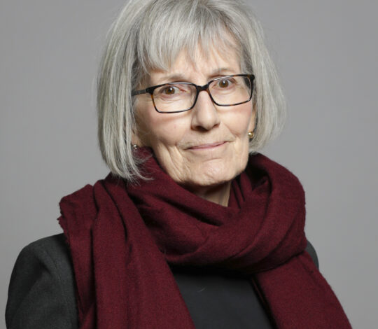 An elderly woman with gray hair wearing glasses and a burgundy scarf stands against a gray background.