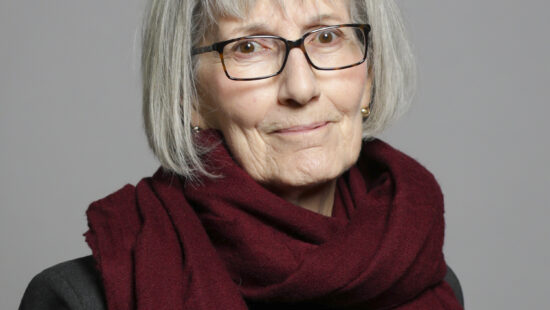 An elderly woman with gray hair wearing glasses and a burgundy scarf stands against a gray background.