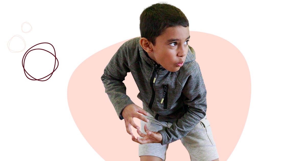 A young boy in a grey hoodie and light shorts crouches in a playful stance against a pink and black background with doodle circles.