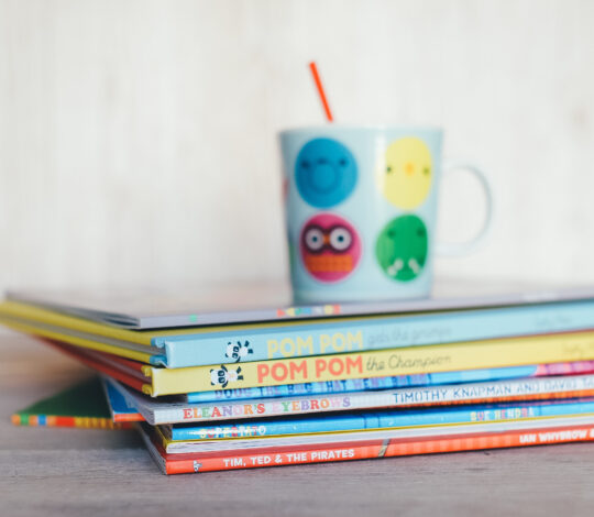 A stack of colorful children's books with a cartoon-themed coffee cup on top, placed on a wooden surface.