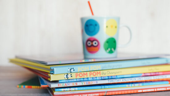 A stack of colorful children's books with a cartoon-themed coffee cup on top, placed on a wooden surface.