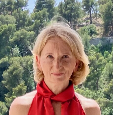 A woman with short blonde hair in a red halter top stands outdoors with a background of green trees and hills.