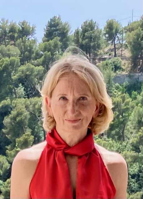 A woman with short blonde hair in a red halter top stands outdoors with a background of green trees and hills.