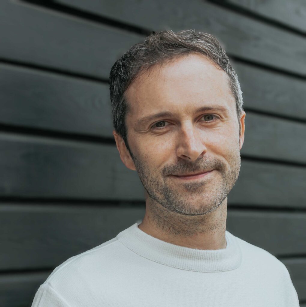 A man with short hair wearing a white sweater with colorful horizontal stripes stands against a dark wooden wall, looking at the camera and slightly smiling.