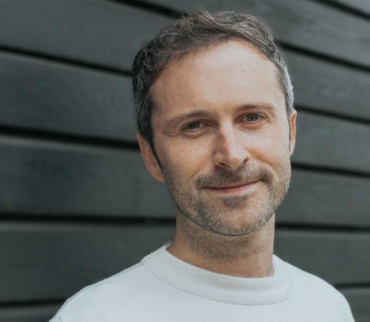 A man with short hair wearing a white sweater with colorful horizontal stripes stands against a dark wooden wall, looking at the camera and slightly smiling.